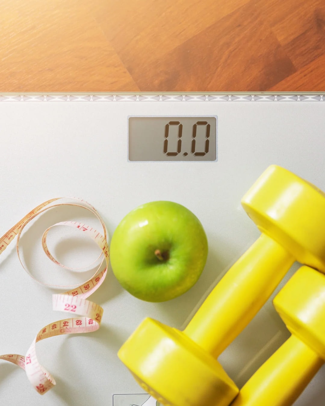 Bathroom scales reading zero beside an apple, a tape measure and a pair of yellow dumbbells