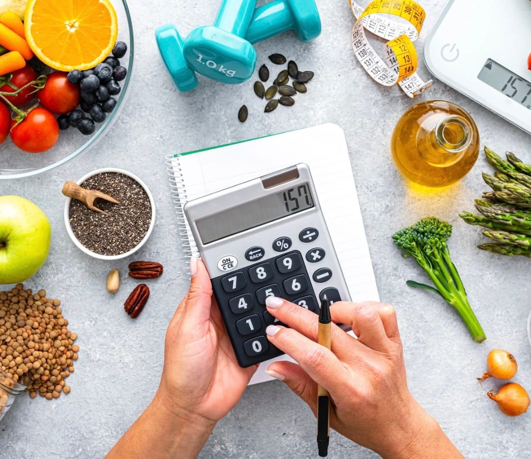 Hands using a calculator surrounded by fruit, vegetables, seeds, oil and kitchen scales