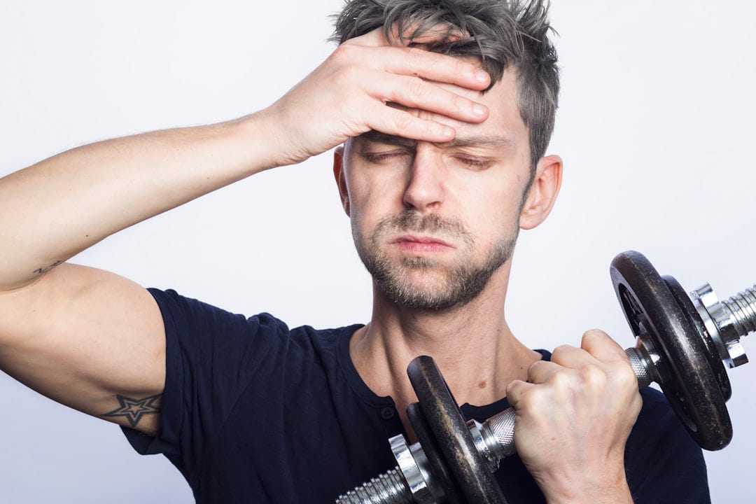 A man holding a dumbbell with one hand pressed to his forehead, looking exhausted