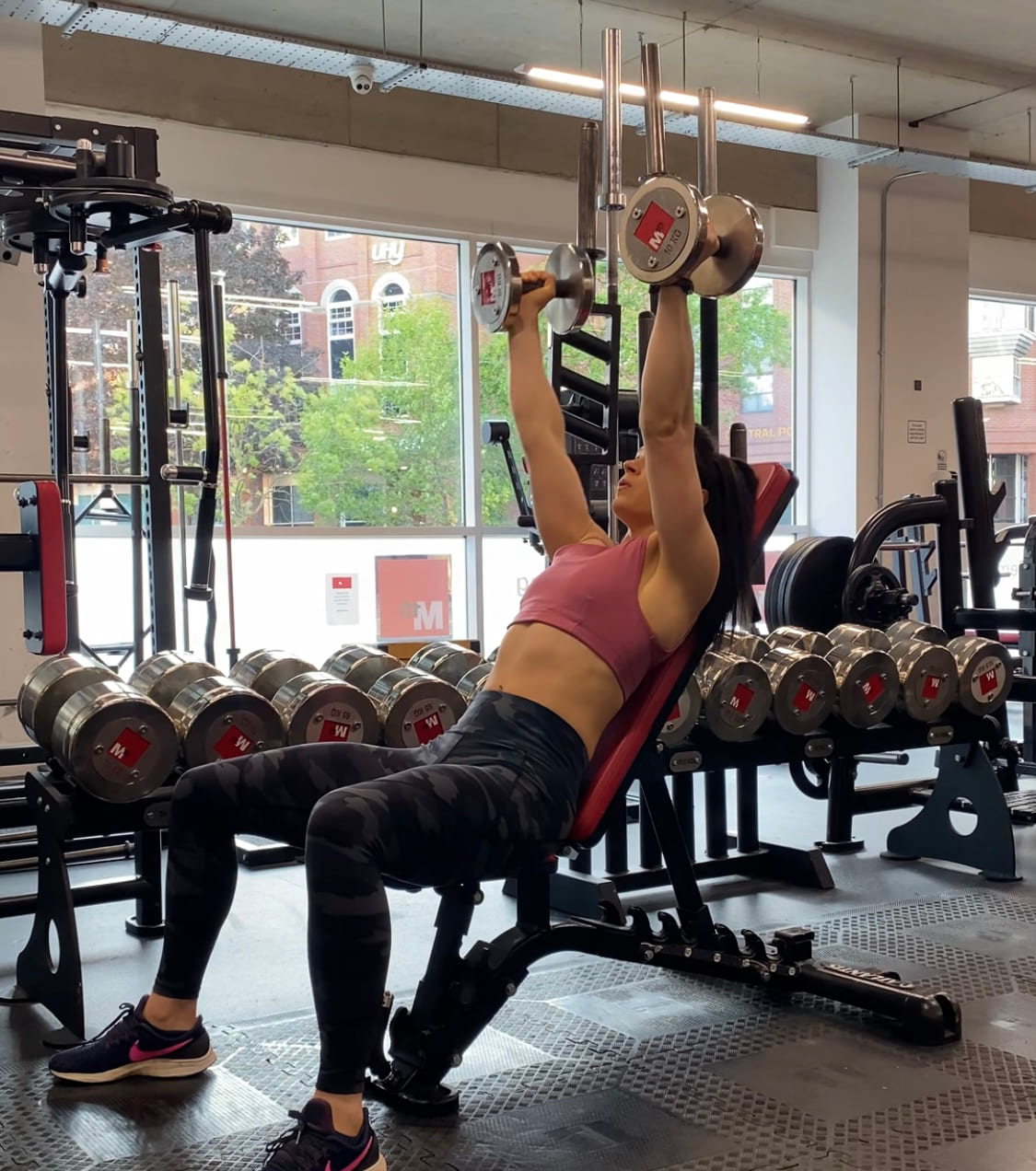 A woman pressing dumbbells overhead on an incline bench beside a window