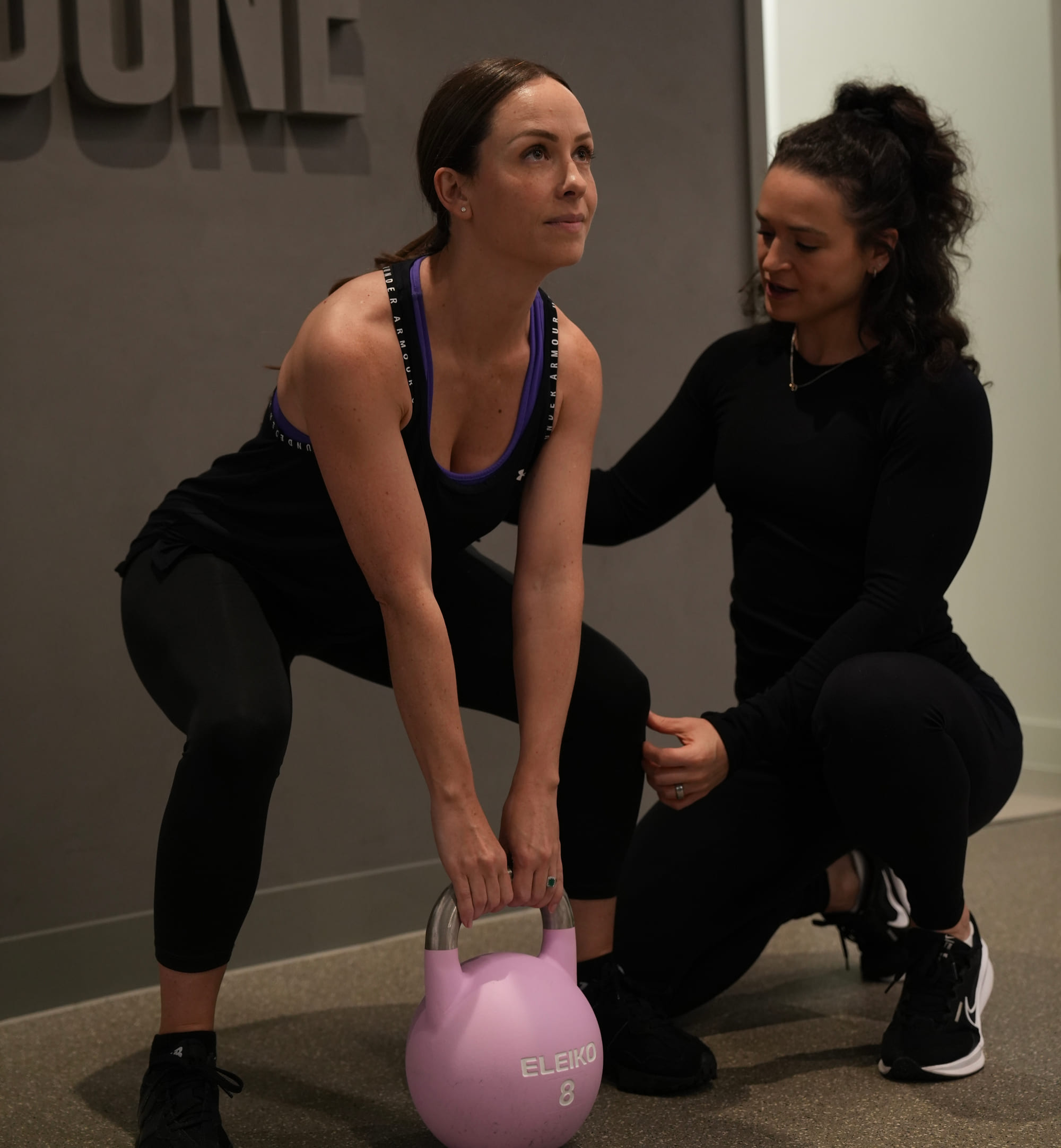 A coach crouching beside a woman lifting a kettlebell from the floor in a training studio