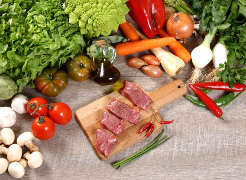 Raw beef on a wooden board surrounded by vegetables, herbs and mushrooms