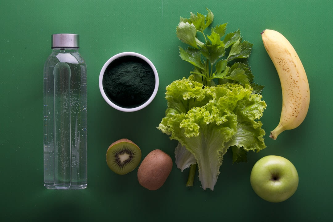 A water bottle, a bowl of green powder, lettuce, a banana, a kiwi and an apple on a green background