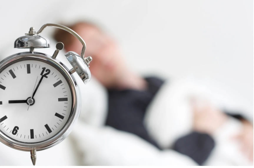 An alarm clock in focus with a sleeping person blurred behind it