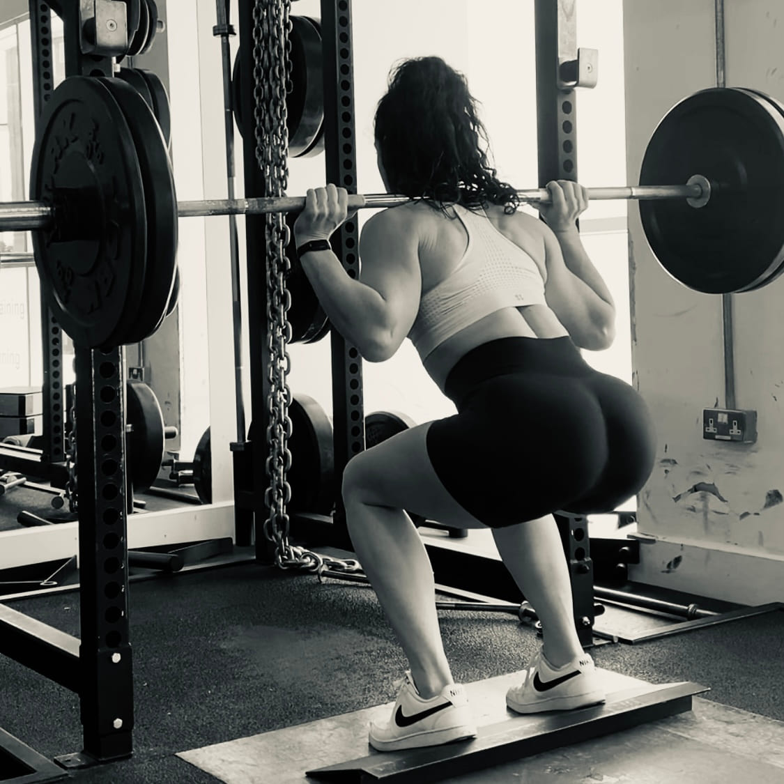 A black and white photograph of a woman squatting with a barbell in a rack