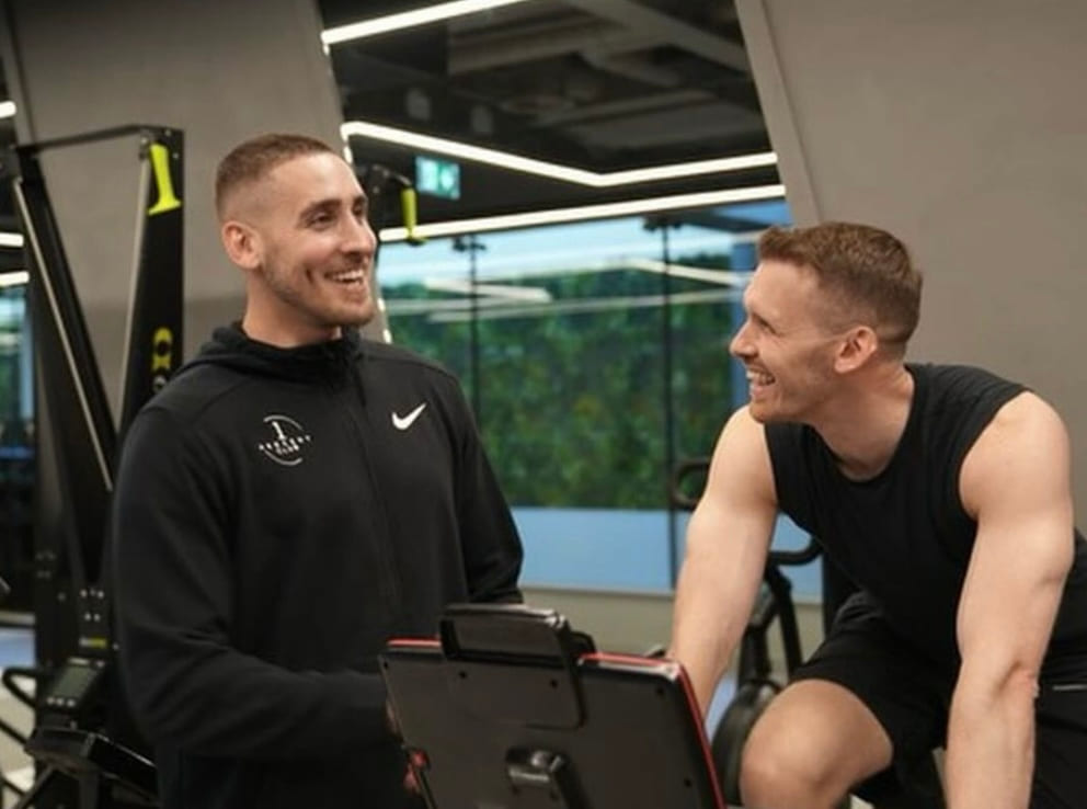 Two men talking beside gym equipment, one holding a clipboard