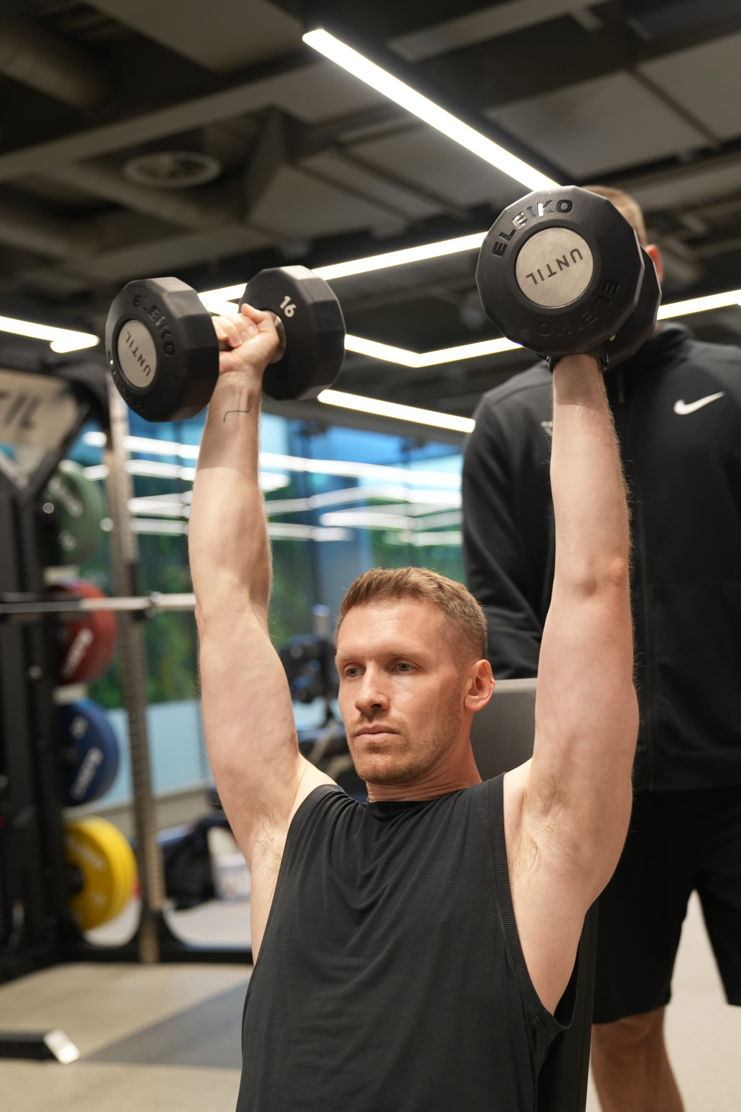 A client pressing dumbbells overhead with a coach spotting