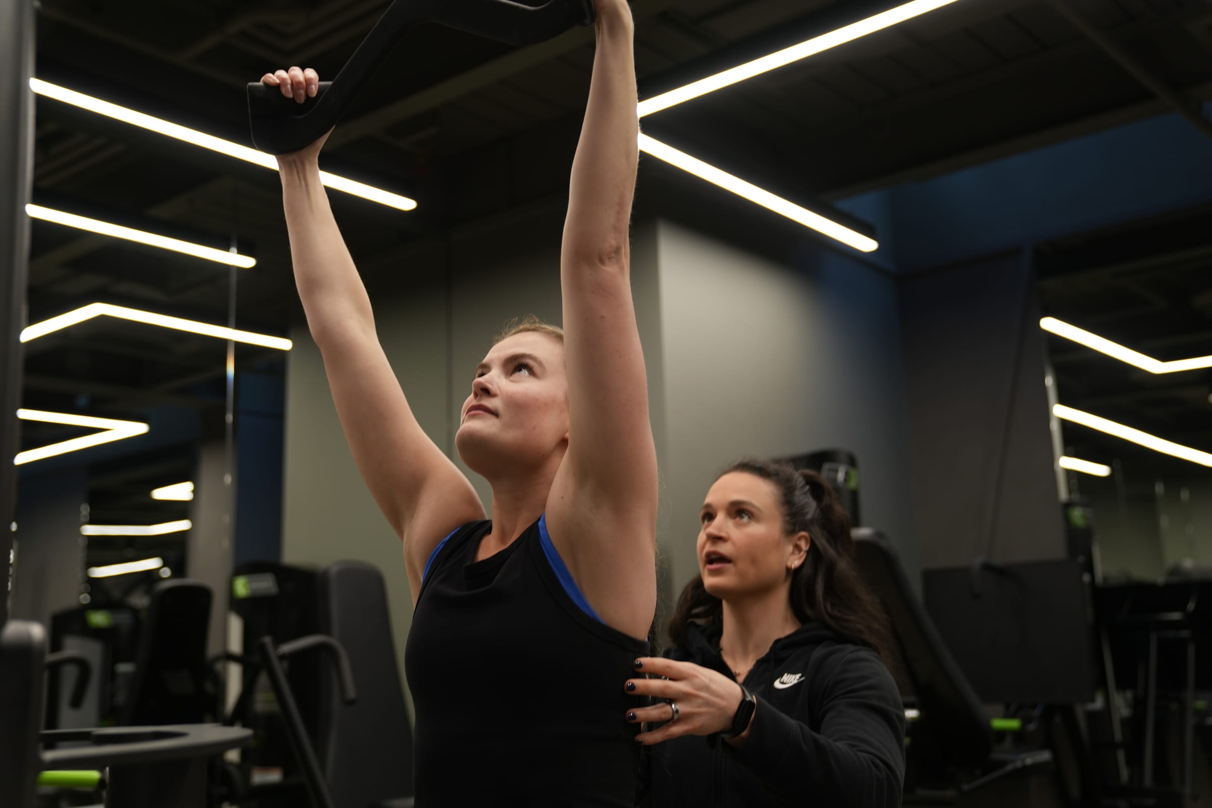 A coach guiding a client through an overhead reach on the cable machine